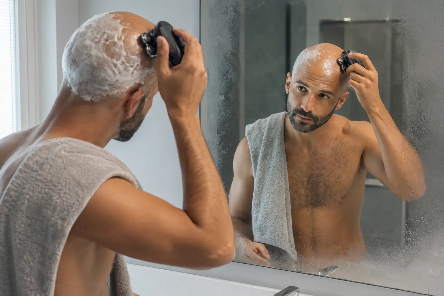 Hombre afeitándose la cabeza con una afeitadora rotativa frente al espejo